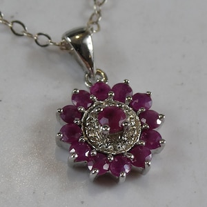 May include: A silver pendant necklace featuring a flower-shaped design with a central round ruby surrounded by a halo of small white stones and an outer ring of twelve round rubies, all set in a polished silver metal frame on a delicate chain.