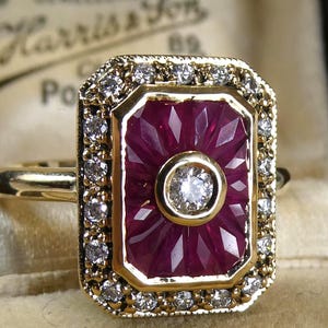 Könnte beinhalten: Goldener Ring mit einem rechteckigen, tiefroten Rubin-Mittelstein, umgeben von einem Kranz aus kleinen Diamanten. Der Rubin ist facettiert, und in der Mitte befindet sich ein einzelner Diamant. Vintage-Stil.
