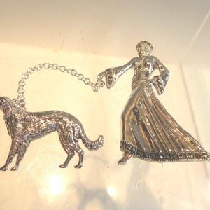 May include: A silver brooch depicting a woman in a long dress holding a dog on a chain. The brooch is decorated with marcasite stones.