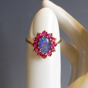 Może przedstawiać: Złoty pierścionek z owalnym, niebieskim opalem otoczonym małymi, czerwonymi kamieniami. Pierścionek jest prezentowany na białej powierzchni, podkreślając szczegóły biżuterii. Kamienie tworzą kwiatowy wzór.