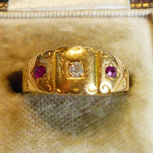 Puede incluir: Un anillo de oro con un diamante y dos rubíes. El anillo está en una caja de terciopelo.