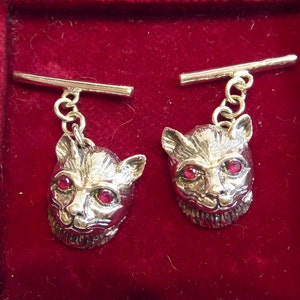 May include: A pair of silver cufflinks featuring a cat head design. Each cufflink has a detailed cat head with red gemstone eyes and a silver bar.
