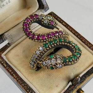 May include: A double-headed snake ring featuring a dark metal band encrusted with round-cut red rubies, green emeralds, and clear white diamonds. The serpent heads are positioned at opposite ends, showcasing intricate bead-set gemstone detailing.