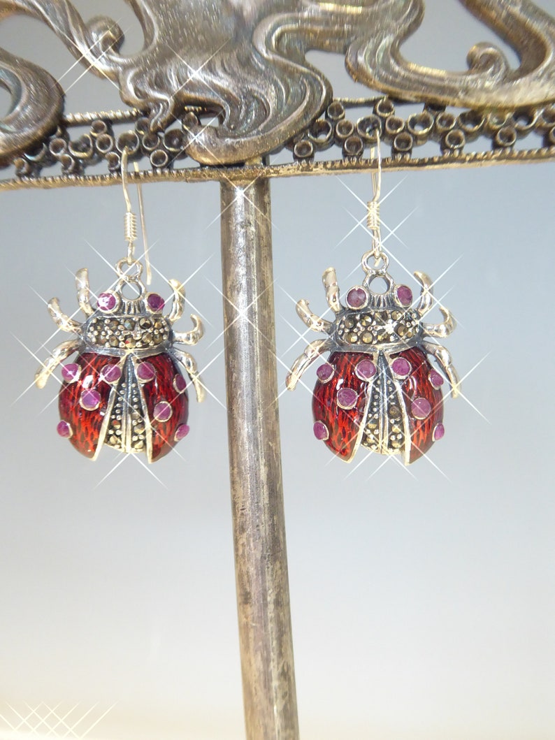 May include: A pair of silver earrings with red enamel ladybug designs. Each ladybug is decorated with small pink gemstones and black marcasite stones.