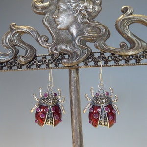 May include: A pair of silver earrings with red enamel ladybug designs. Each ladybug has a black and white marbled pattern and is accented with small pink gemstones.