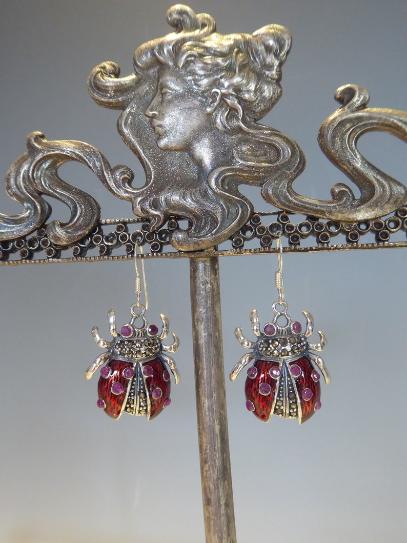 May include: A pair of silver earrings with red enamel ladybug designs. Each ladybug has a black and white marbled pattern and is accented with small pink gemstones.