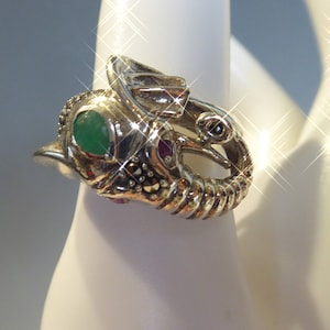 Peut inclure: Bague en argent avec un motif d'éléphant orné de pierres précieuses vertes et rouges. La trompe de l'éléphant est enroulée et ses yeux sont sertis de pierres noires.