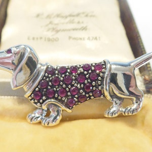 May include: A silver dachshund dog brooch, adorned with numerous small, round, deep red gemstones. The brooch is presented in a vintage jewellery box with a cream-coloured interior.