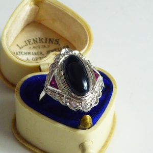 May include: A vintage silver ring with an oval black onyx stone, surrounded by small diamonds and two red stones, set in a heart-shaped jewelry box with blue velvet lining. The box is cream-colored and has the text "L. JENKINS, WATCHMAKER, JEWELER & OPT."