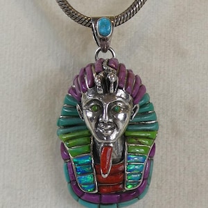 May include: Silver pendant necklace featuring a detailed pharaoh design. The pendant has a turquoise stone at the top and is adorned with colorful stones in shades of purple, green, and red. The pharaoh's face is silver with green eyes.