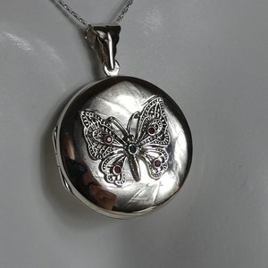 May include: Silver locket necklace featuring a butterfly design. The butterfly has red gemstone accents.