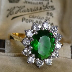 Può includere: Un anello d'oro con una grande pietra verde ovale al centro, circondata da un gruppo di pietre più piccole e trasparenti. L'anello è posto su uno sfondo color crema, con il testo "H. Harris & Son" visibile.