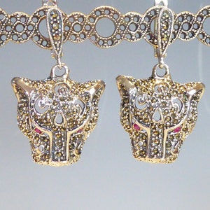 May include: A pair of silver earrings featuring a panther head design. The panther heads are crafted from filigree and decorated with marcasite stones. The earrings have a red stone for the eyes.