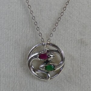 May include: A silver pendant necklace featuring two intertwined snakes forming a circular shape. One snake head is set with an oval-cut deep red gemstone, while the other snake head is set with an oval-cut vibrant green gemstone. The pendant hangs from a delicate silver chain.