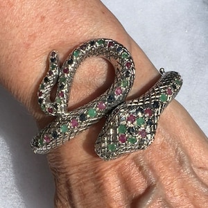 Puede incluir: Una pulsera de serpiente plateada con un diseño texturizado en forma de escamas. El cuerpo de la serpiente está adornado con piedras preciosas rojas, verdes y azules. La pulsera se envuelve alrededor de la muñeca, creando un accesorio llamativo.