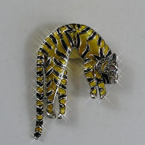 Puede incluir: Broche de tigre esmaltado en amarillo y negro con marco de metal plateado. El broche presenta un diseño detallado de un tigre curvado, con una cabeza adornada con pedrería.
