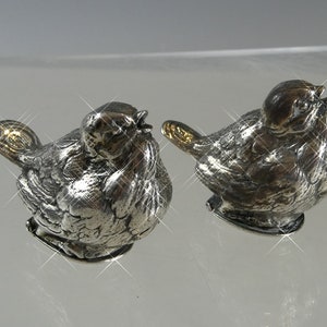 Pode incluir: Duas estatuetas de pássaros de prata com detalhes dourados. Os pássaros estão de frente um para o outro e têm detalhes elaborados.