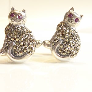 May include: A pair of silver cat earrings with red gemstone eyes and a marigold-colored stone body.