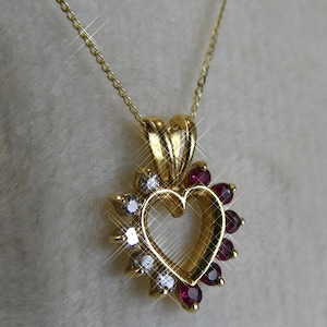 Może przedstawiać: Złoty naszyjnik z wisiorkiem w kształcie serca. Serce otoczone jest naprzemiennie przezroczystymi i czerwonymi kamieniami szlachetnymi. Łańcuszek naszyjnika jest również złoty, tworząc spójny i elegancki design.