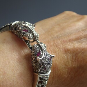 May include: Silver bracelet with two jaguar head motifs, adorned with ruby and black gemstones. The bracelet incorporates marcasite stones and detailed patterns. A piece of jewellery.