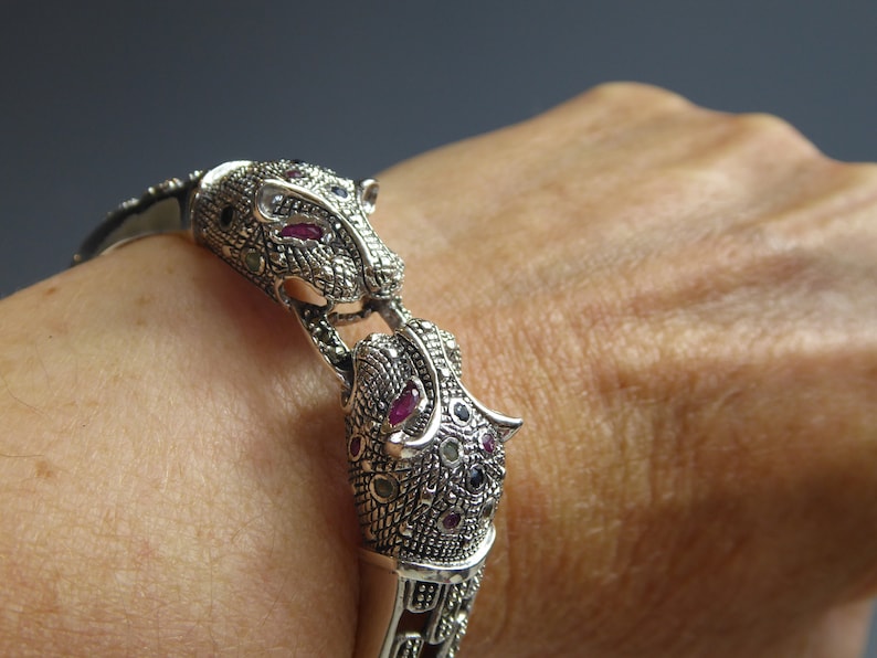 May include: Silver bracelet with two jaguar head motifs, adorned with ruby and black gemstones. The bracelet incorporates marcasite stones and detailed patterns. A piece of jewellery.