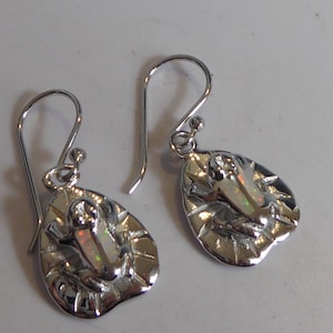 Puede incluir: Un par de pendientes de plata con un diseño de rana. Cada rana está sentada en una hoja y tiene una piedra de ópalo blanco en su cuerpo.