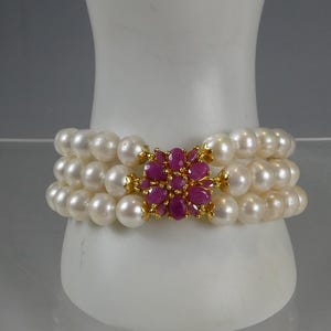 Pode incluir: Uma pulseira de pérolas de três fios com um fecho dourado com um aglomerado de pedras preciosas de rubi. As pérolas são brancas e redondas, e os rubis são de um vermelho profundo. A pulseira é exibida em uma superfície branca.