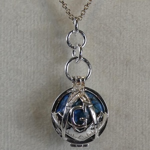 Könnte beinhalten: Silberfarbene Anhängerkette mit einem kugelförmigen Anhänger mit blauem Stein. Der Anhänger ist in einem dekorativen Käfigdesign mit dem Buchstaben "G" eingefasst. Die Kette hat eine feine Kette. Der Durchmesser beträgt ca. 2,5 cm.