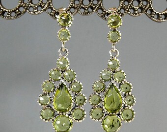 May include: A pair of dangling earrings with a silver-tone metal setting. Each earring features a cluster of round and teardrop-shaped, light green gemstones. The earrings are suspended from a decorative, circular metal bar.