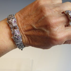May include: Silver-toned bracelet and ring set with a jaguar design. The bracelet has a hinged clasp and is decorated with red, blue, and clear gemstones. The matching ring features a jaguar head with similar gemstone detailing.