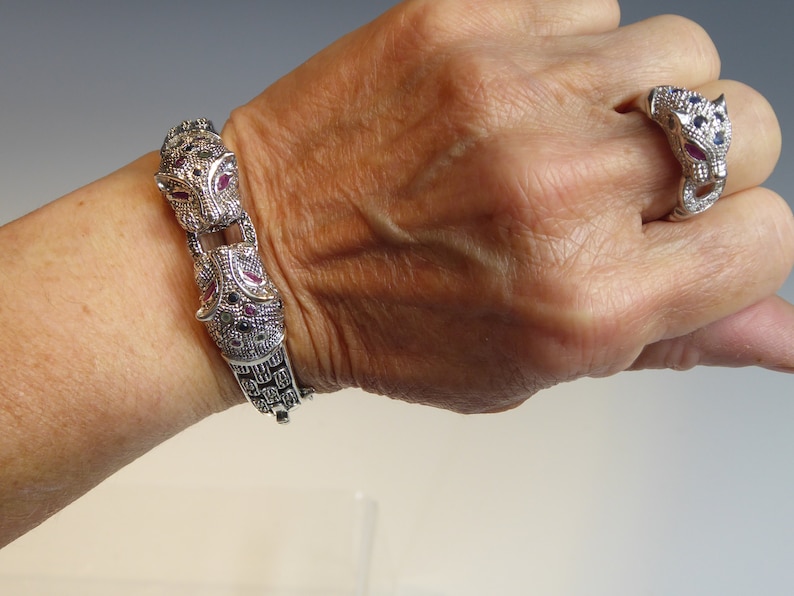 May include: Silver-toned bracelet and ring set with a jaguar design. The bracelet has a hinged clasp and is decorated with red, blue, and clear gemstones. The matching ring features a jaguar head with similar gemstone detailing.