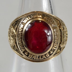 May include: Gold-toned class ring with a large, oval, red gemstone center. The ring features detailed engravings, including text that reads "MORROW WILSON SCHOOL DALLAS 1970". The ring is set against a clear background.