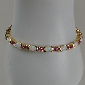 Puede incluir: Un brazalete de oro con ópalos ovalados alternados y pequeños rubíes redondos. Las piedras preciosas están engastadas en una banda de oro. El brazalete se exhibe sobre una superficie blanca.