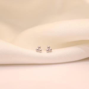 May include: A pair of small, silver stud earrings, each with three tiny, spherical beads arranged in a triangular cluster. The earrings are displayed on a white, textured fabric background.