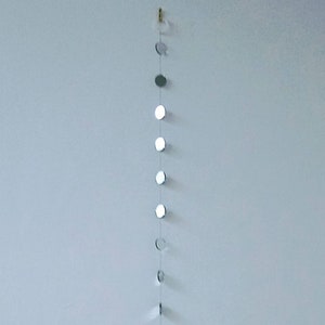 May include: A decorative wall hanging featuring a series of small, circular mirrors arranged in a vertical line. The mirrors reflect light, creating a shimmering effect. The piece is suspended from a small hook, adding a touch of elegance to any space.
