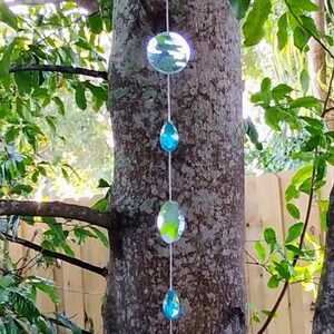 May include: A decorative hanging ornament with clear and blue glass elements. The ornament is suspended from a string and attached to a tree trunk. The design includes round and teardrop-shaped glass pieces.