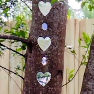 May include: A decorative heart-shaped wind chime hanging from a tree. The chime features multiple heart-shaped and geometric-shaped reflective pieces, along with clear crystal accents. The tree's bark is brown, and green leaves and branches surround the chime.