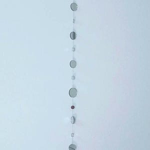 May include: A decorative hanging mirror garland. The garland features several small, round, reflective mirrors of varying sizes strung together with clear string. The mirrors are silver and hang vertically, creating a shimmering effect. The garland is suspended from a clear loop.