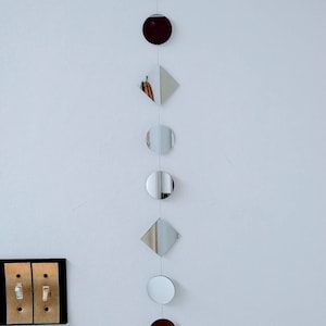 May include: A decorative wall hanging with reflective circles and diamond shapes, strung together with a clear string. A crystal prism hangs at the bottom. The wall hanging is mounted on a white wall. The shapes are in various sizes.