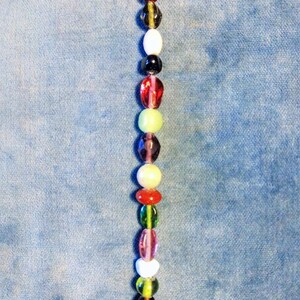 May include: A vertical string of assorted beads in a variety of colours and shapes. The beads include red, green, yellow, white, and purple hues. The beads are strung on a silver-coloured wire, creating a decorative accessory.