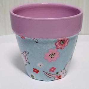 May include: A small flower pot with a solid light pink painted rim and a light blue fabric covering on the base. The fabric features a repeating pattern of small pink flowers and white rabbit illustrations scattered across the light blue background.