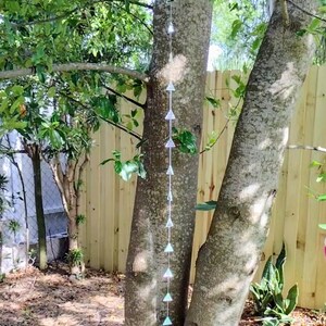 May include: A decorative outdoor hanging chain with triangular prism-shaped ornaments. The chain is suspended from a tree branch, with the ornaments reflecting light. The background includes trees, foliage, and a wooden fence.