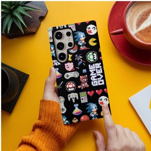May include: Black phone case with a pixelated video game design featuring characters like Sonic the Hedgehog, mushrooms, and game controllers. The case has the words "GAME OVER" in white.