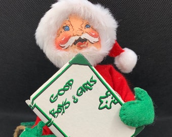 Könnte beinhalten: Eine Plüsch-Weihnachtsmann-Puppe hält ein Buch mit der Aufschrift "Good Boys & Girls". Er trägt einen roten Anzug, einen weißen Bart und grüne Fäustlinge. Er sitzt auf einer grünen Oberfläche mit einer verpackten Geschenkbox.