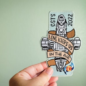 May include: A blue and white embroidered patch featuring a spaceship with the text "LOVE KEEPS US IN THE AIR" on a banner and "CSTS 2022" on the side.