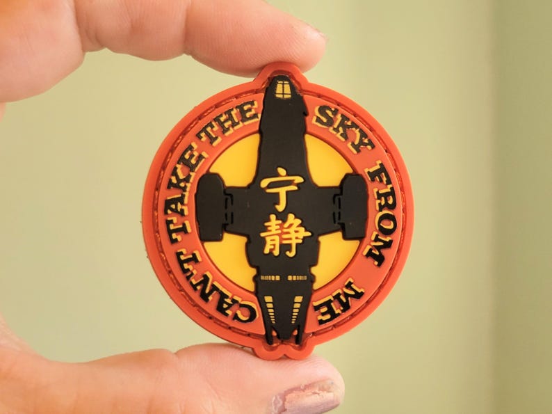May include: A round, orange and yellow patch with the text "CAN'T TAKE THE SKY FROM ME" encircling a black spaceship design. The center features Chinese characters. A collectible item.