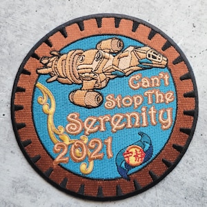 Op de afbeelding: Een ronde, geborduurde patch met een bruine buitenring en een blauw centrum. De patch heeft een ruimteschip, de tekst "Can't Stop The Serenity 2021" en een klein blauw en rood symbool.