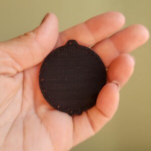 May include: A dark brown, circular patch with a textured surface and a small loop at the top. The patch is held in a hand against a soft green background. The patch appears to be made of a fabric material.