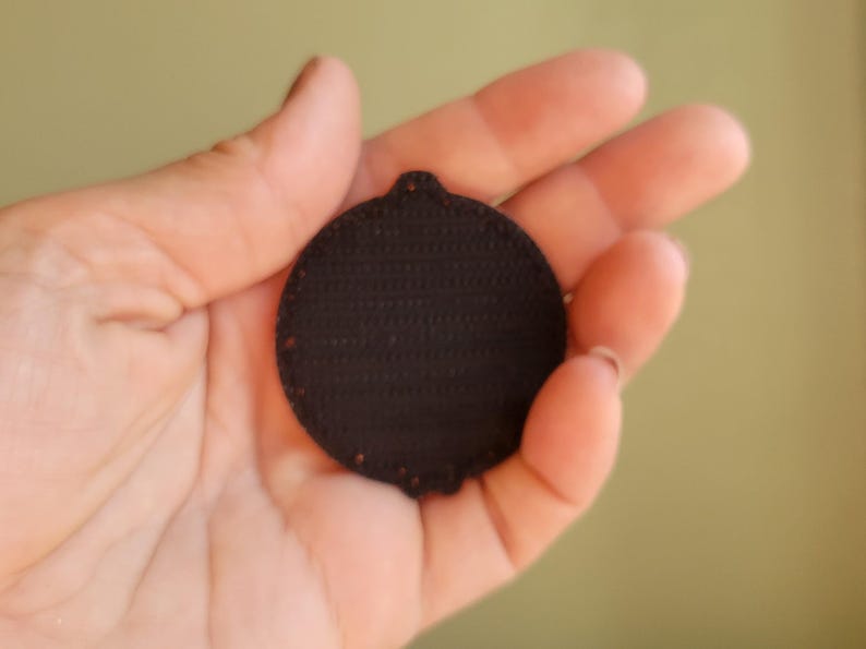 May include: A dark brown, circular patch with a textured surface and a small loop at the top. The patch is held in a hand against a soft green background. The patch appears to be made of a fabric material.
