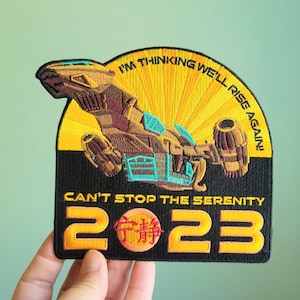 May include: A black and yellow embroidered patch featuring a spaceship with the text "I'm thinking we'll rise again! Can't stop the Serenity 2023".